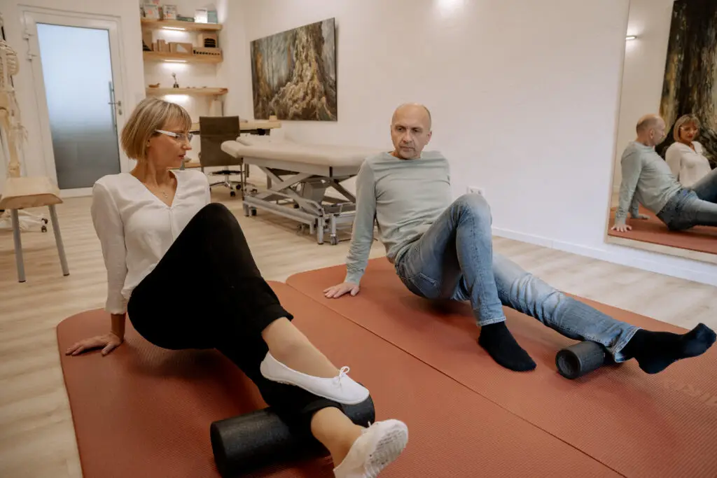 Gabriele Cunskis zeigt einem Patienten auf der Übungsmatte die Faszienrollmassage.